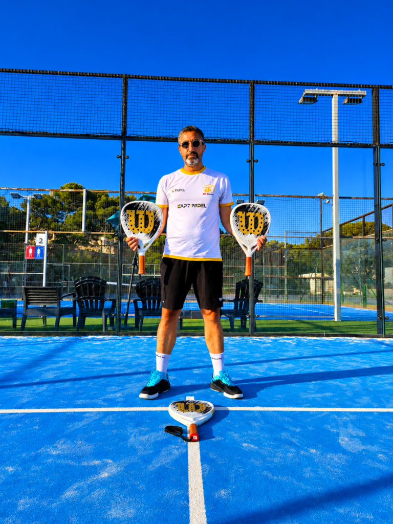 Adult padel player in a white jersey and black shorts stands on a bright blue court, holding two paddles with a paddle on the ground between his feet.