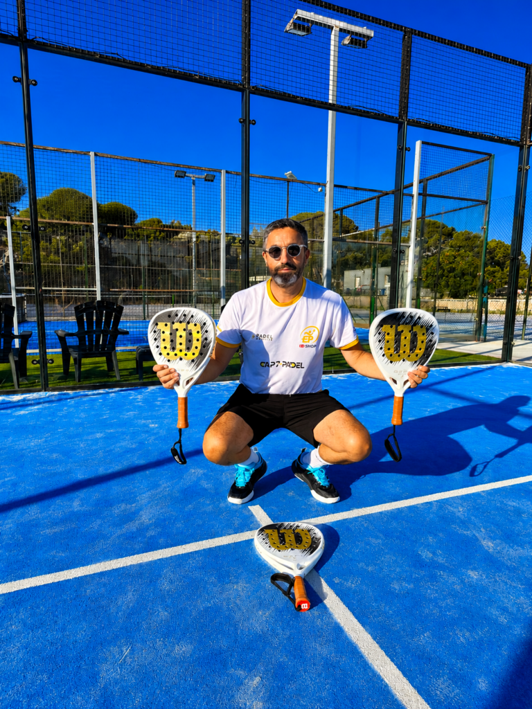 Man on a blue padel court squats and holds two white rackets with orange handles, with a third racket on the ground behind him.