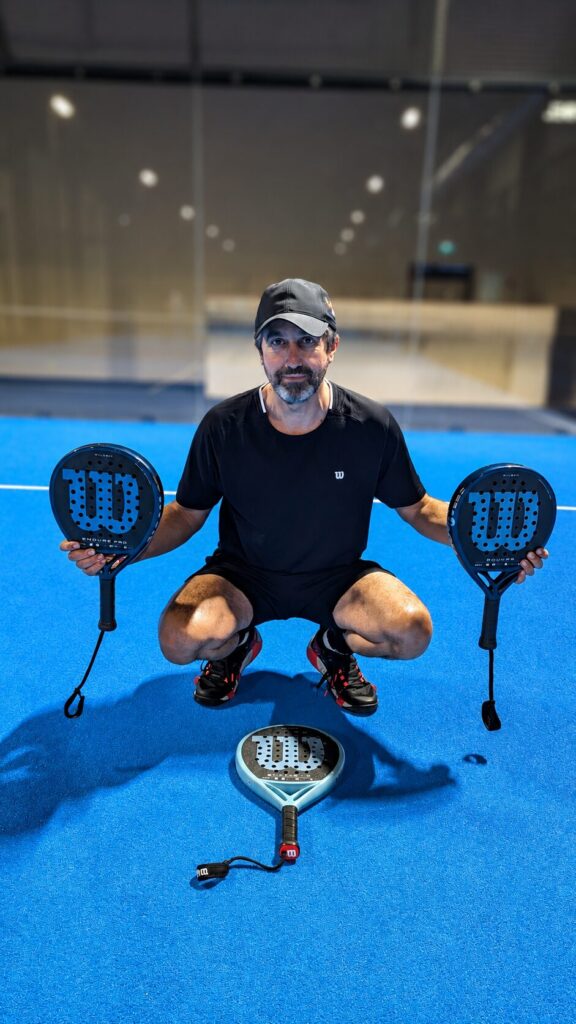 Man kneeling on a blue padel court, holding two padel rackets, with a racket on the floor in front of him.