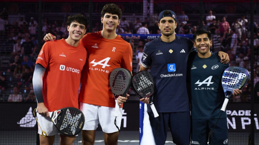 Four male padel players stand side by side on court, holding paddles, smiling as spectators watch in the background.