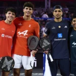Four male padel players stand side by side on court, holding paddles, smiling as spectators watch in the background.