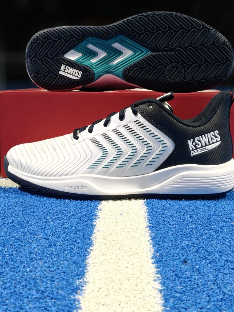 oplus_2162736 | Padel Magazine White and black K-Swiss athletic shoe with a blue patterned outsole, resting on a red box against a blue track with a white line.