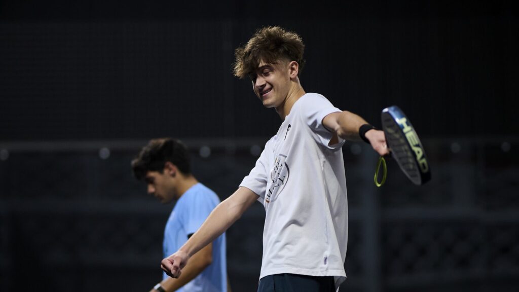 Curro Cabeza, 18 ans et déjà 49e mondial : le « Roi Arthur » n’a qu’à bien se tenir | Padel Magazine A young man in a white shirt swings a tennis racket indoors, smiling as he hits the ball.
