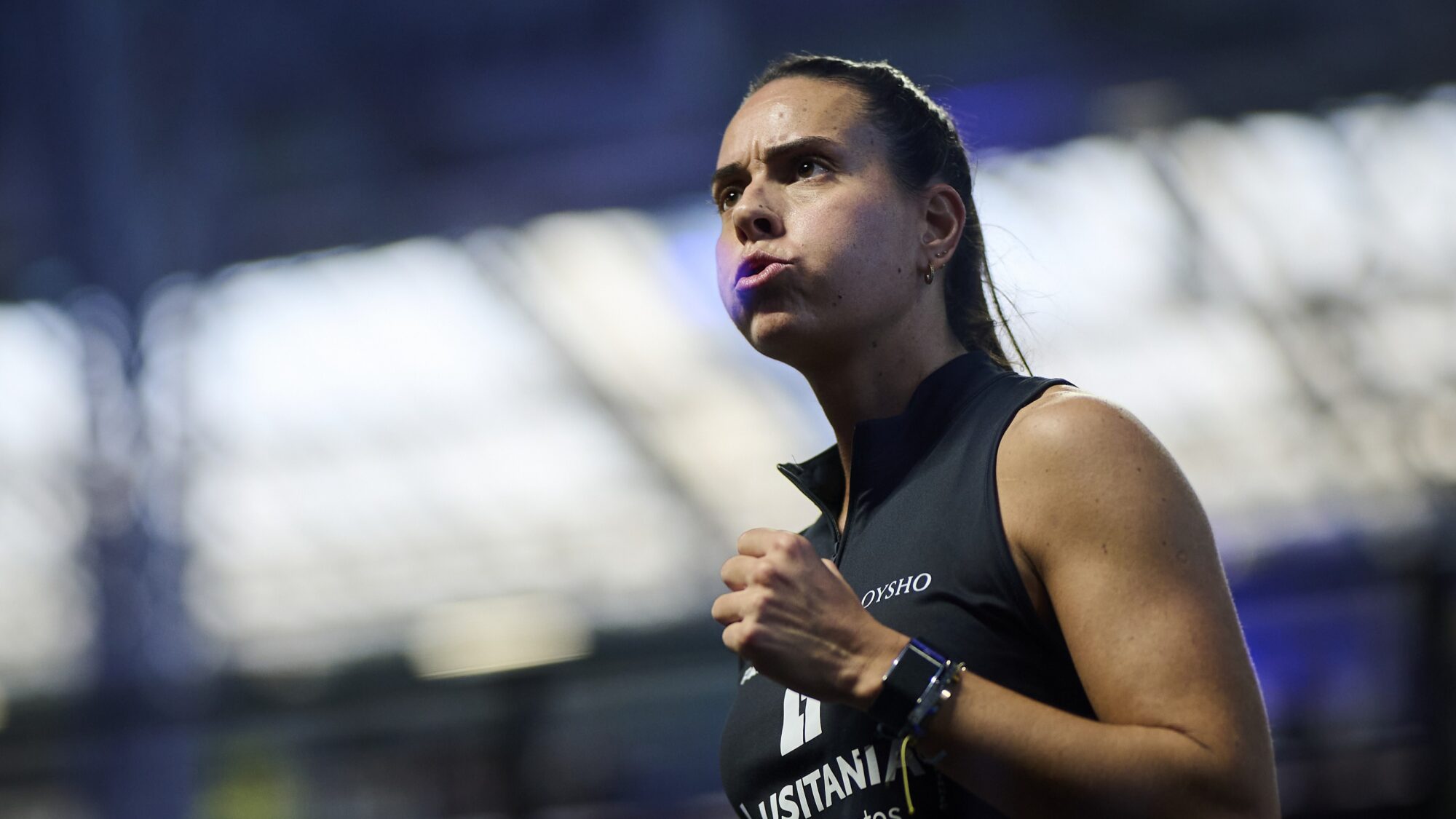 Female runner in a sleeveless black top, mid-stride with a focused expression, bib number visible on her chest in a stadium setting.