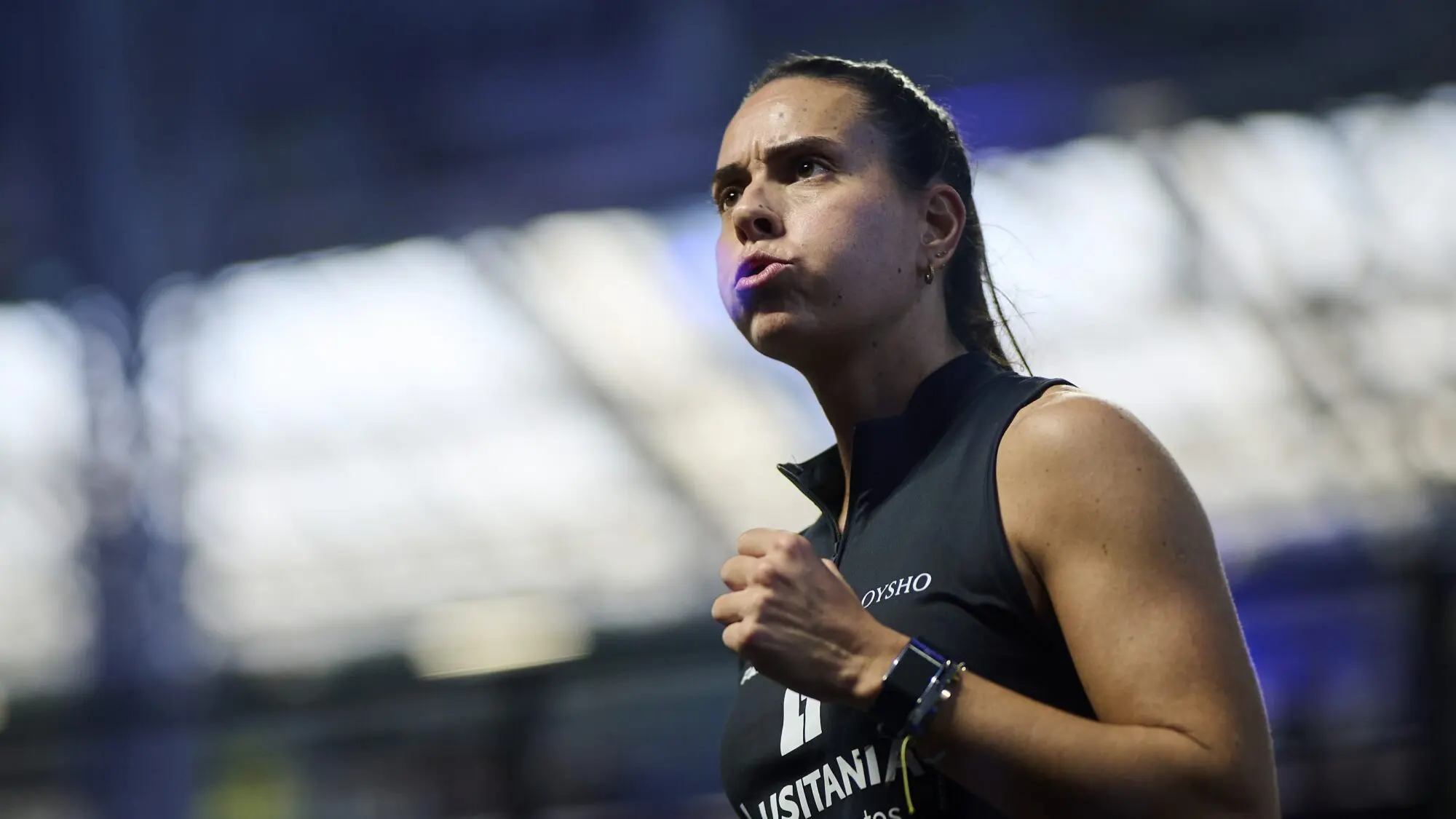 Female runner in a sleeveless black top, mid-stride with a focused expression, bib number visible on her chest in a stadium setting.