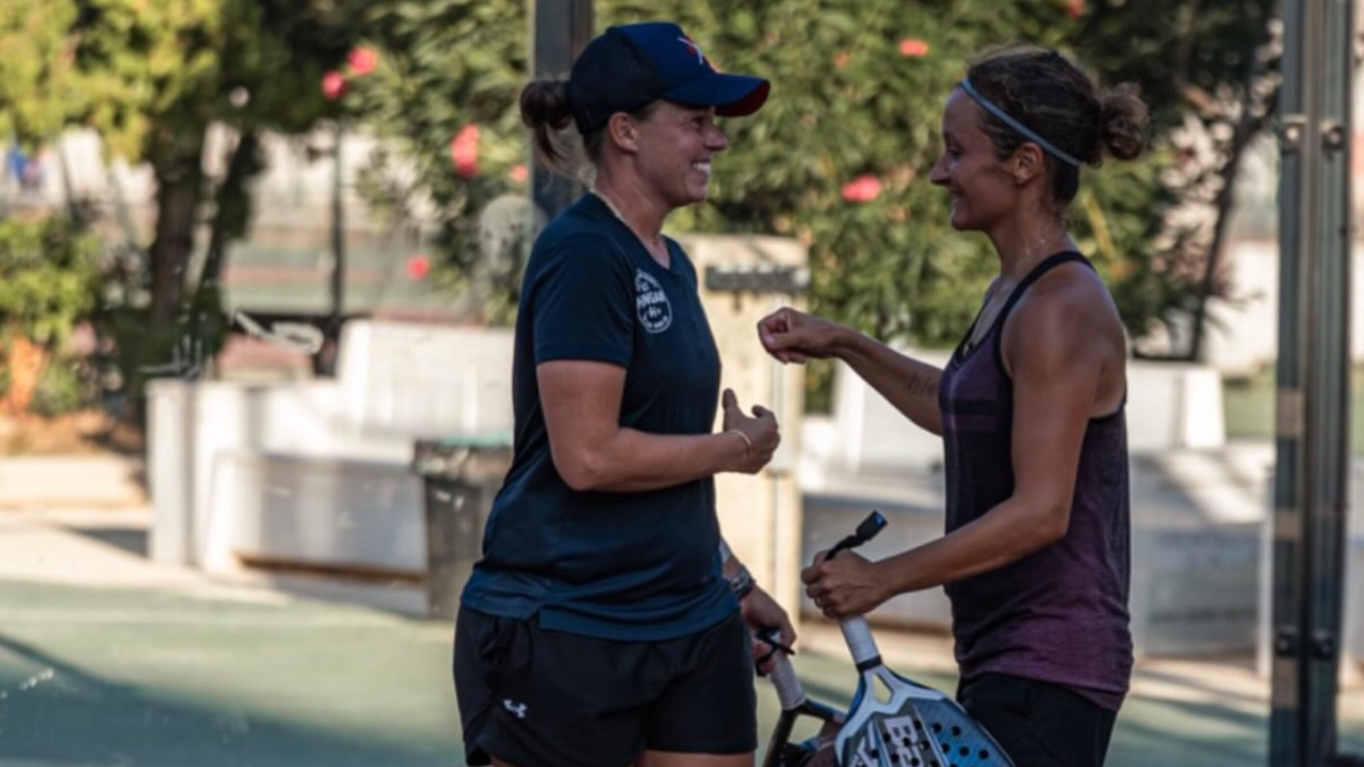 Two women in athletic wear share a fist bump outdoors near a sports court, smiling.