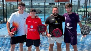 Four male padel players pose together on a blue court, each holding a paddle and smiling at the camera.