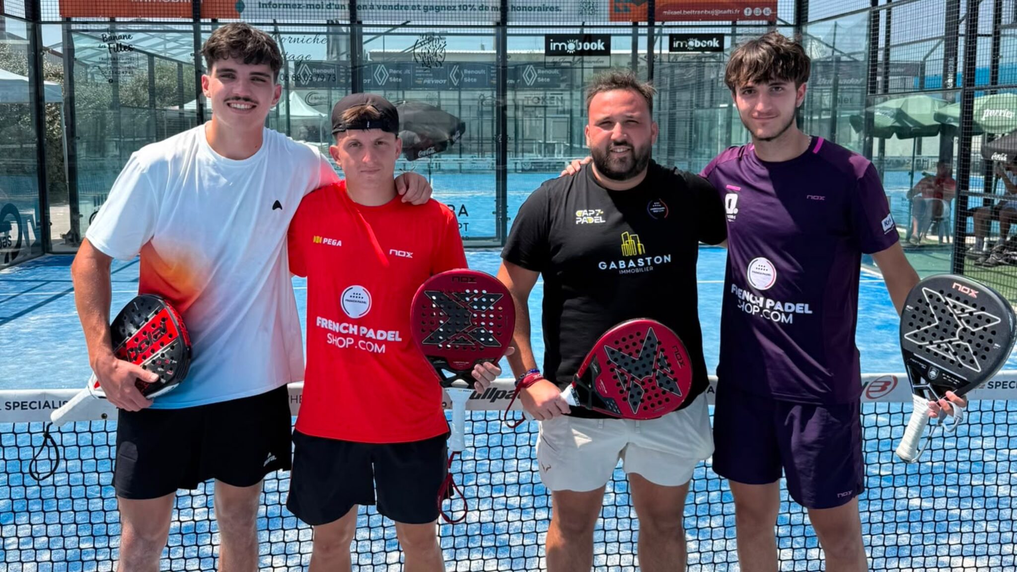 Four male padel players pose together on a blue court, each holding a paddle and smiling at the camera.
