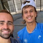Blanqué et Joris en finale du FIP Bronze de Frascati, une première ensemble | Padel Magazine Two men smiling for a selfie at a padel court, with a chain-link fence in the background; one wears a blue shirt and a backward white cap.
