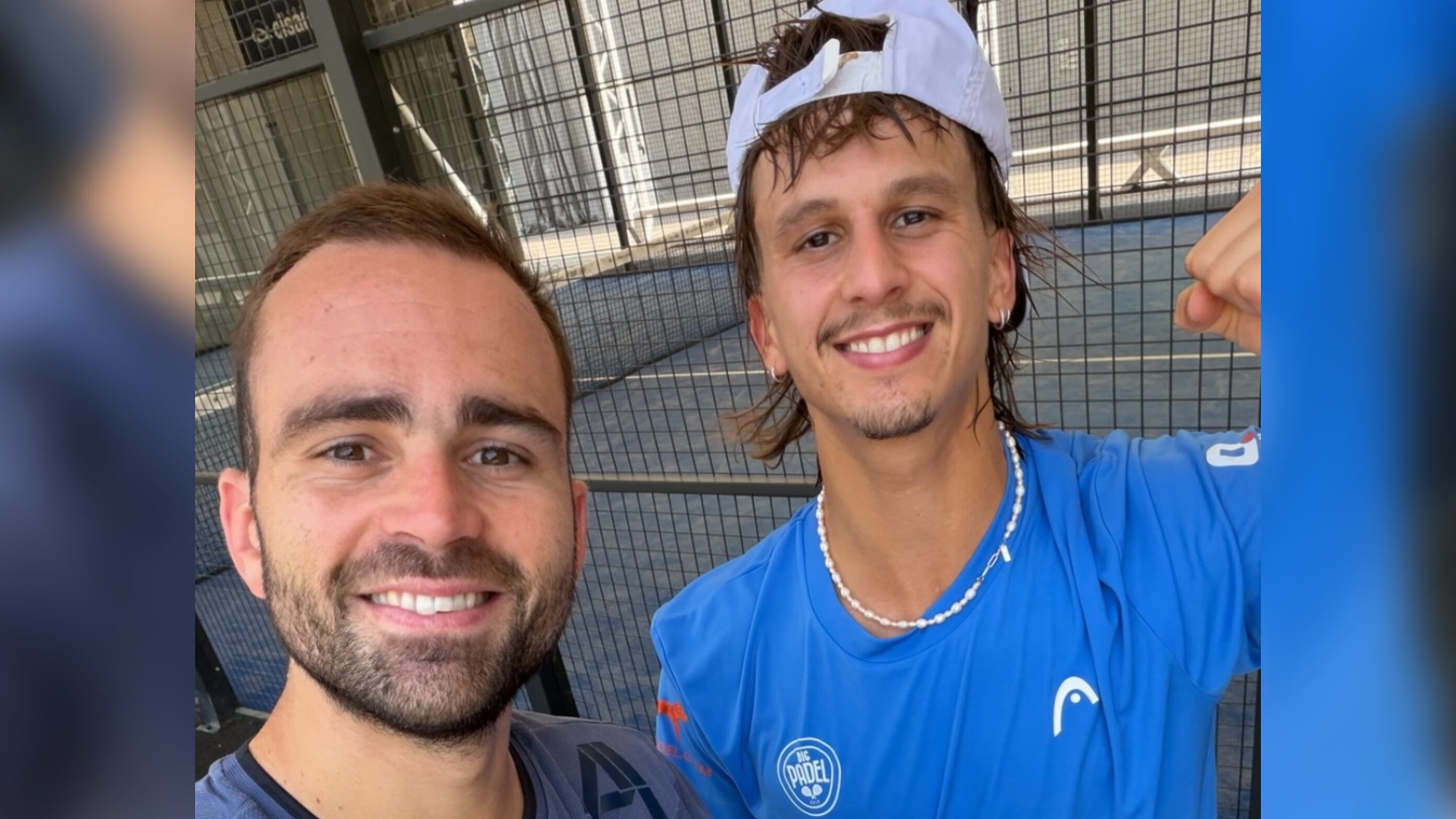 Two men smiling for a selfie at a padel court, with a chain-link fence in the background; one wears a blue shirt and a backward white cap.