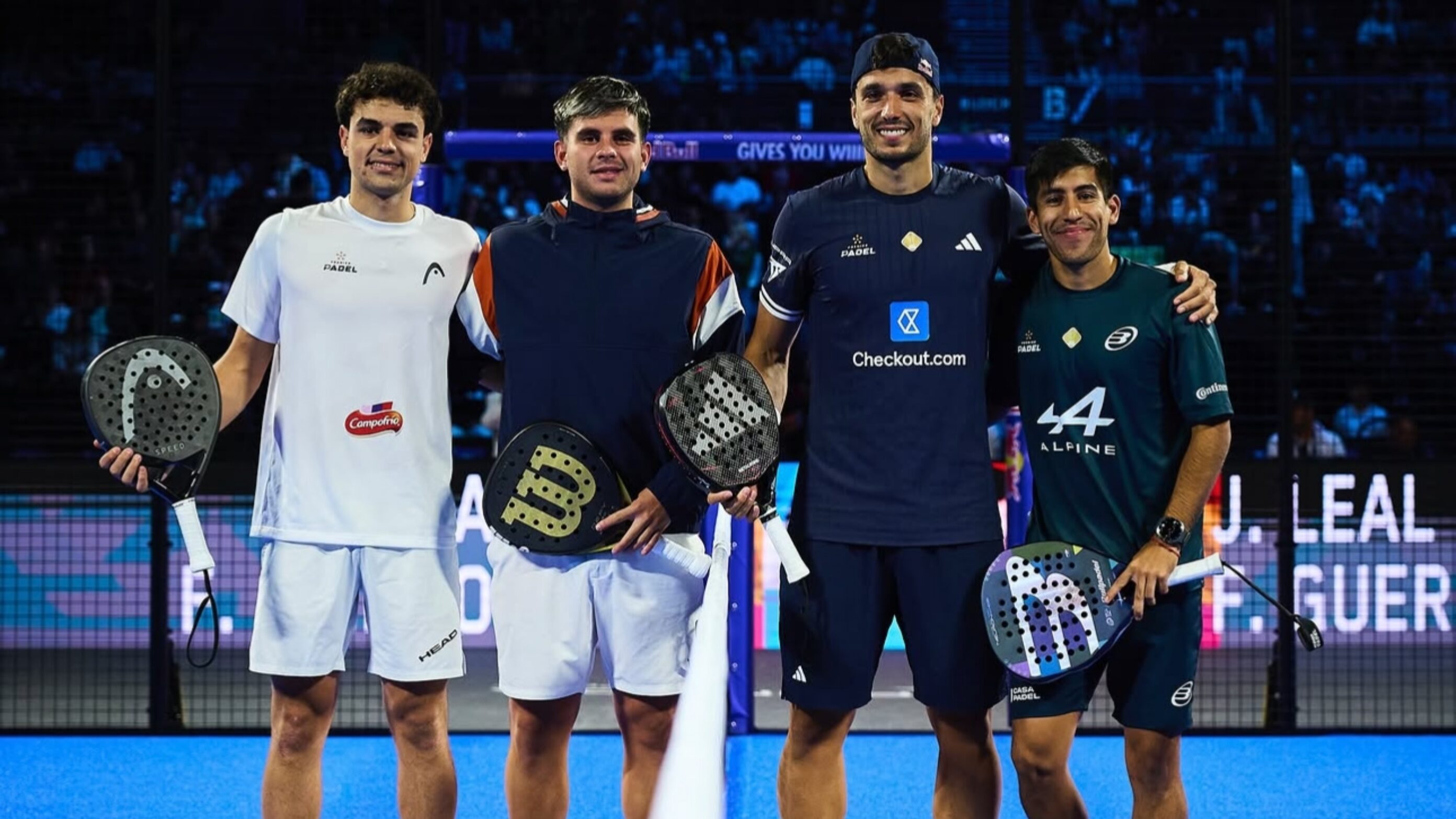 Four male padel players stand together on a blue court, holding their paddles and smiling after a match.