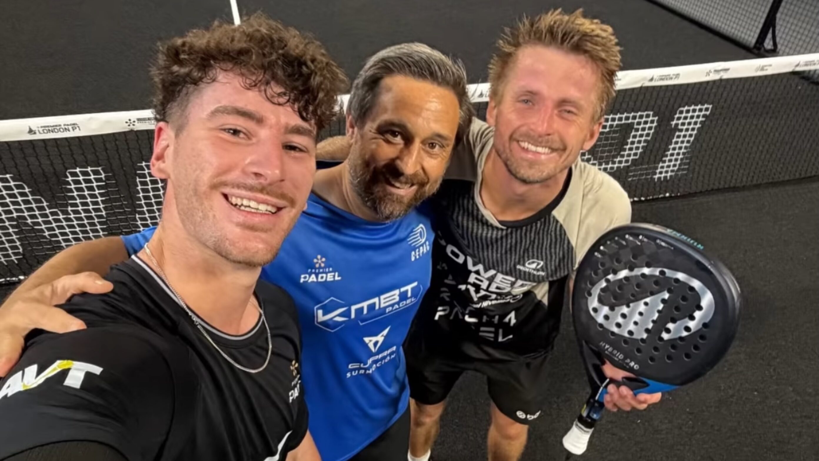 Three men in sports gear taking a selfie on a padel court, smiling with a paddle visible in the foregroundrocking a close-knit group vibe