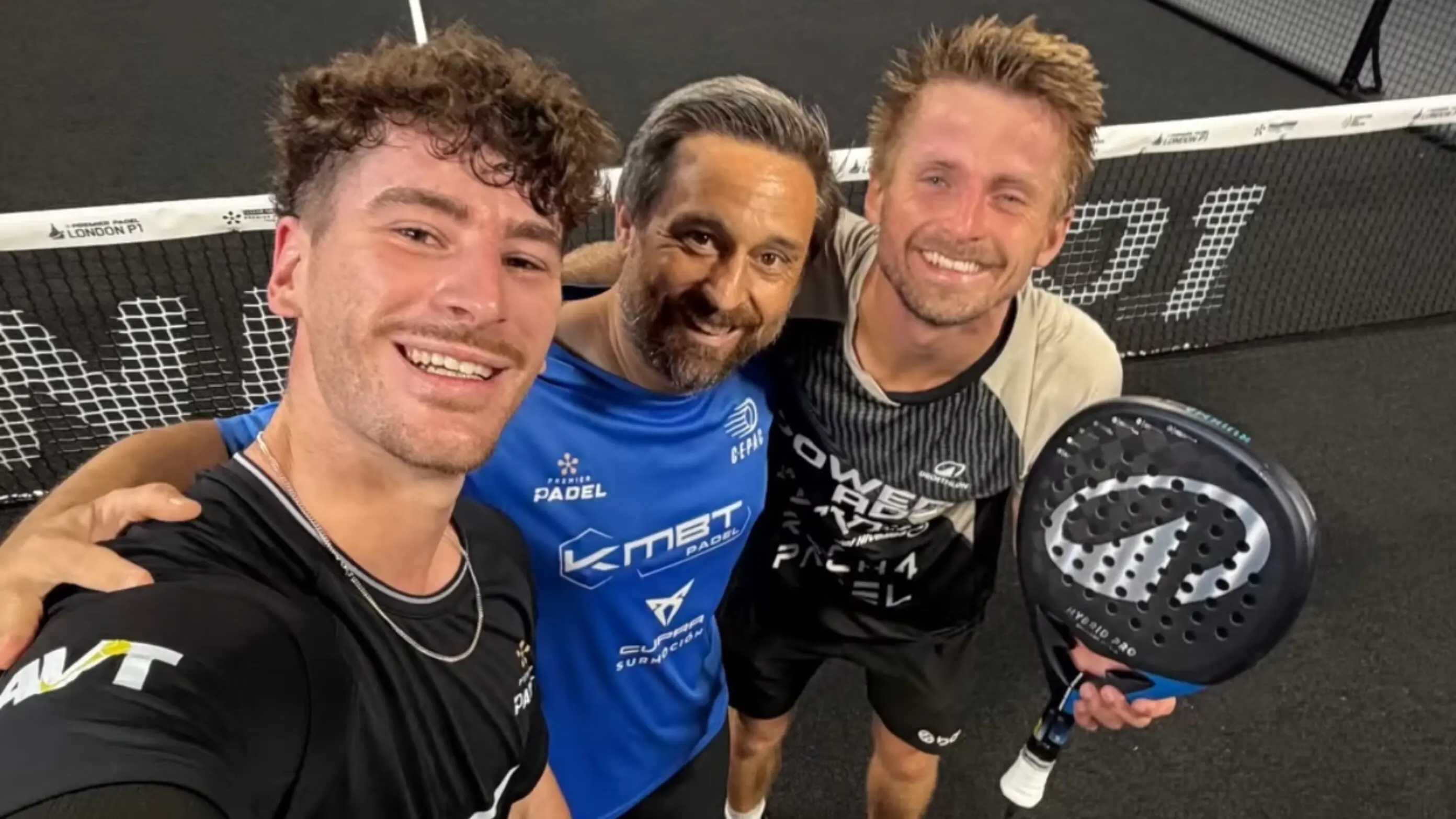 Three men in sports gear taking a selfie on a padel court, smiling with a paddle visible in the foregroundrocking a close-knit group vibe