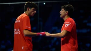 Two male athletes in orange jerseys shake hands on an indoor court after a match.