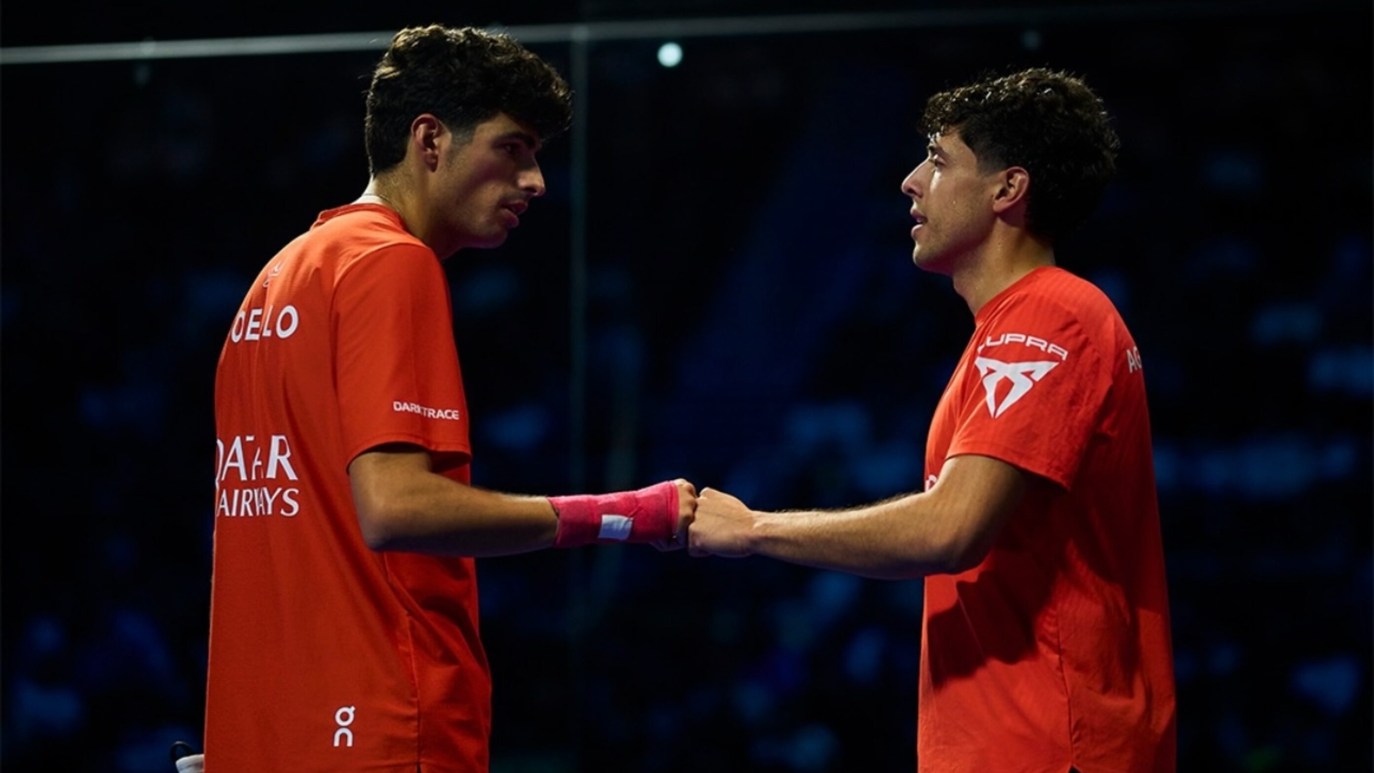 Two male athletes in orange jerseys shake hands on an indoor court after a match.