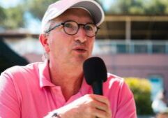 Man in a pink polo and white cap speaks into a handheld microphone outdoors, wearing glasses.