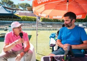 Two men interview outdoors on a sunny sports field under a large red umbrella; one in a pink polo speaks into a handheld mic, the other in a blue shirt wears a headset.