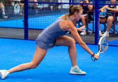 Female padel player in a blue outfit lunges to strike a ball with a perforated paddle on a bright blue court, behind a blue fence with spectators.