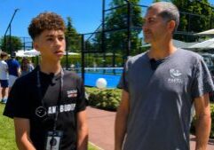 Two men stand near a blue paddle tennis court on a sunny day; the younger wears a black shirt with a lanyard and mic, the older in a gray'Padel Magazine' shirt listening.