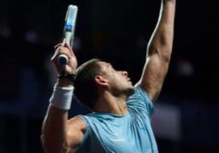 Athlete in a blue shirt raises arms in celebration during a padel match, holding a paddle racket in one hand.