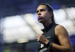 Female runner in a sleeveless black top, mid-stride with a focused expression, bib number visible on her chest in a stadium setting.